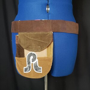 May include: A brown corduroy belt with a patchwork pouch. The pouch features various shades of brown corduroy and a white sticker with a stylized "R" design. The belt is displayed on a blue mannequin.