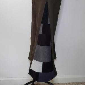May include: Olive green pants featuring a side slit that reveals a patchwork panel. The panel consists of rectangular fabric segments in shades of black, charcoal gray, and light gray, arranged in a grid pattern along the lower leg section.