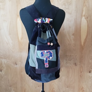 May include: A patchwork backpack with a drawstring closure, featuring a variety of fabrics in shades of blue, green, and black. A pocket displays a large question mark with red stars. The top has a colorful yin-yang pattern.
