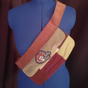 May include: A patchwork crossbody bag in shades of red, tan, and olive green. The bag features a wide strap and a decorative patch with a skull design. The bag is displayed on a blue mannequin.
