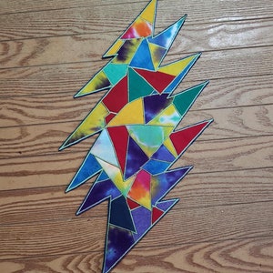 May include: A colorful, tie-dye lightning bolt design with a black outline. The bolt is composed of various geometric shapes in shades of red, yellow, blue, green, and purple. The background is a wooden surface.