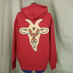 May include: Red hooded sweatshirt with a cream-colored goat graphic on the back. The goat design features brown and teal accents. The sweatshirt has a hood and long sleeves.