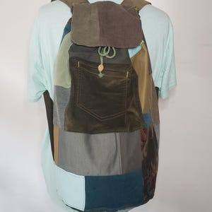 May include: A patchwork backpack constructed from various fabric panels in shades of olive green, brown, teal, and light blue. The bag features a top flap, a drawstring closure with a green cord and wooden bead, and a large front pocket.