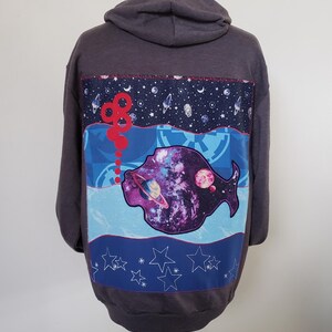 May include: Dark gray hoodie with a colorful space-themed design on the back. The design features a fish shape with a galaxy inside, planets, stars, and a red, looping design. The hoodie has a hood and long sleeves.
