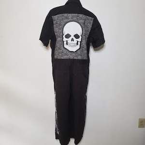 May include: A black short-sleeved jumpsuit featuring a large square patch on the back with a white skull graphic set against a black and white swirling pattern. The garment has a collar, a button-up front, and visible stitching along the legs.