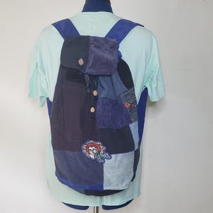 May include: A patchwork backpack constructed from various shades of blue fabric, including corduroy and denim. The bag features a flap closure with two wooden buttons, blue shoulder straps, and a circular embroidered patch depicting a skull.