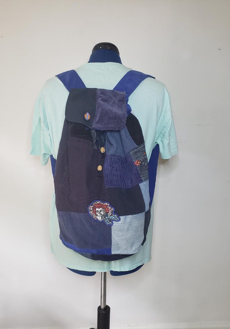 Może przedstawiać: Plecak typu patchwork wykonany z r&oacute;żnych odcieni niebieskiego materiału, w tym sztruksu i dżinsu. Torba posiada klapę z dwoma drewnianymi guzikami, niebieskie szelki oraz okrągłą, haftowaną naszywkę przedstawiającą czaszkę.