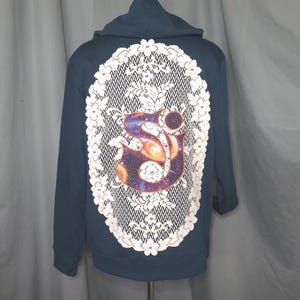 May include: A dark teal hoodie with a white lace design on the back. The lace frames a colorful graphic of a galaxy-like design with the letter "S" in the center. The hoodie has a hood and long sleeves.