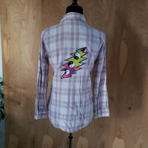 May include: A light pink and gray plaid button-down shirt with long sleeves. The back of the shirt features a colorful lightning bolt design with yin-yang symbols. The shirt is displayed on a mannequin.