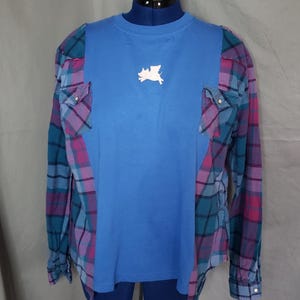 May include: A blue long-sleeve shirt with plaid sleeves and accents. The shirt features a white pig graphic on the front. The plaid incorporates teal, purple, and pink colors. Two pockets are visible on the sleeves.