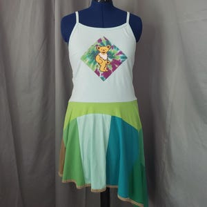 May include: A light blue tank top dress with a colorful bear graphic on the front. The dress features a patchwork skirt in shades of green, blue, and brown. The dress is displayed on a mannequin.