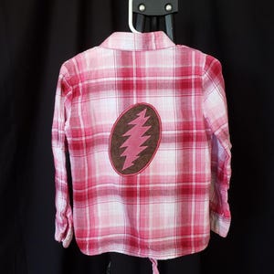 May include: Pink and white plaid button-down shirt with a brown and pink lightning bolt design on the back. The shirt is hanging on a white hanger against a black background. The shirt has long sleeves.