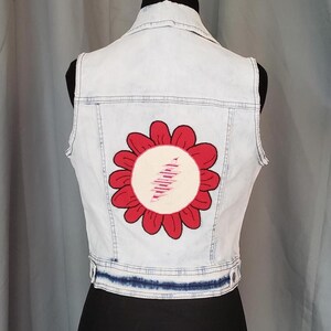 May include: Light blue denim vest with a large red and white flower design on the back. The vest has a classic collar, a button-up front, and a distressed look. The flower has a white center with pink lines.