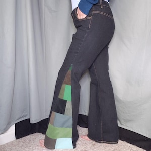 May include: Dark wash denim jeans with a flared leg. The left leg features a patchwork design with squares of brown, green, and blue fabrics. The jeans have a classic five-pocket design and a dark brown stitching.