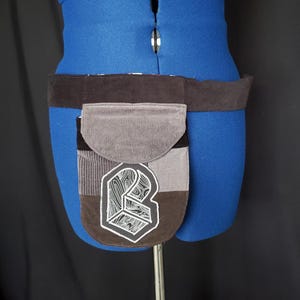 May include: A brown and gray corduroy belt bag with a geometric design. The bag features a flap closure and a patch with a white and black graphic. The belt is dark brown and the bag is attached to a blue mannequin.