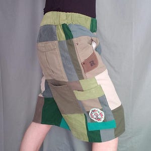May include: Patchwork skirt in shades of green, tan, and blue. The skirt features a variety of fabric squares sewn together, with pockets and a decorative patch. The waistband is elasticized. The skirt is knee-length.