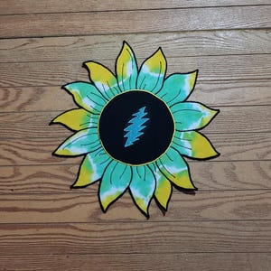 May include: A tie-dye sunflower design with yellow and teal petals, a black center, and a blue lightning bolt symbol. The sunflower is set against a wooden background.