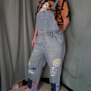 May include: Light blue denim overalls with distressed patches and floral and patterned fabric accents. A large flower patch is on the right leg. The overalls have a bib pocket and adjustable straps. The person is wearing a tie-dye t-shirt with a white graphic.