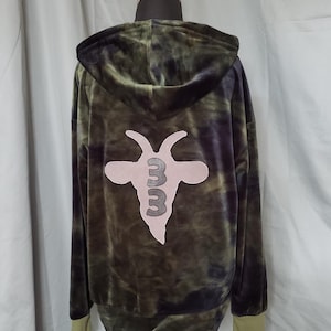 May include: Green and black tie-dye hoodie with a hood and a light pink goat head and "3" graphic on the back. The hoodie has a zip-up front and olive green cuffs. The garment is displayed on a mannequin.