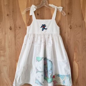 May include: White sundress with tie straps, featuring a blue dancing bear applique on the bodice. The skirt has patchwork panels with whimsical illustrations in pastel colors. The dress is hanging on a clear plastic hanger.