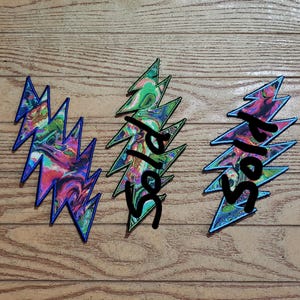 May include: Three lightning bolt shaped fabric patches featuring a vibrant, multi-colored marbled pattern with swirls of pink, blue, green, and purple. The two patches on the right have the word sold written in black across their surfaces.