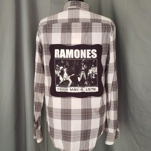 May include: A long-sleeved, button-down shirt with a gray and white plaid pattern. The back of the shirt features a black patch with the word "RAMONES" above a black and white image of the band and the text "CBGB MAY 4, 1978".