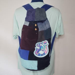 May include: A patchwork backpack constructed from various fabric textures and colors, including dark purple, blue, and light teal panels. The bag features two light-colored wooden buttons and a prominent circular patch with a colorful, swirling graphic design.