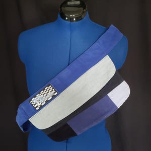 May include: A blue and gray crossbody bag with a checkered flag patch that reads "Family Surfing". The bag is made of various fabric panels in shades of blue, gray, and black. The strap is wide and blue.