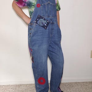 May include: Blue denim overalls featuring a tie-dye t-shirt underneath. The overalls have a rectangular patch with a fish graphic on the chest, two red circular patches on the front, and colorful patterned fabric panels along the side seams.
