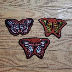 May include: Three butterfly patches with red, black, white, and yellow details are displayed on a wooden surface. Each patch has a unique butterfly design with intricate wing patterns and a black border. The patches are likely for clothing or accessories.