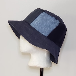 May include: A bucket hat featuring a combination of dark navy blue fabric panels and a contrasting light blue denim-textured square panel on the side. The hat has a wide, downward-sloping brim and a structured crown, displayed on a white mannequin head.