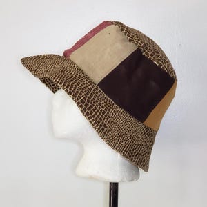 May include: A bucket hat featuring a patchwork design with panels of brown reptile-textured fabric, solid dark brown, tan, and a muted red section. The hat has a wide, downward-sloping brim and a rounded crown, constructed from multiple fabric segments.