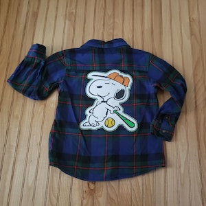 May include: Blue, green, and red plaid button-down shirt featuring a Snoopy baseball graphic on the back. The graphic shows Snoopy holding a baseball bat and a yellow baseball. The shirt has long sleeves.