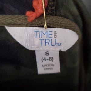 Pode incluir: Close-up de uma etiqueta de roupa de um moletom com capuz. A etiqueta exibe "TIME AND TRU" em letras azuis. Abaixo, indica o tamanho "S (4-6)" e "MADE IN CHINA". Um alfinete de seguran&ccedil;a dourado e linha laranja est&atilde;o anexados.