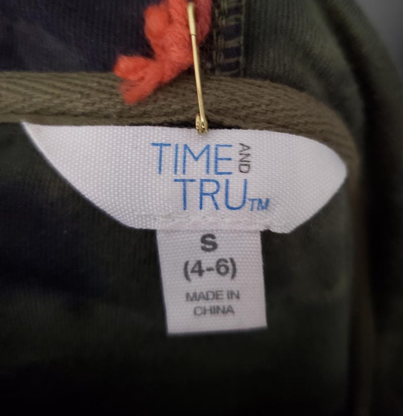 Pode incluir: Close-up de uma etiqueta de roupa de um moletom com capuz. A etiqueta exibe "TIME AND TRU" em letras azuis. Abaixo, indica o tamanho "S (4-6)" e "MADE IN CHINA". Um alfinete de seguran&ccedil;a dourado e linha laranja est&atilde;o anexados.