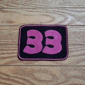 May include: A black rectangular patch with the number "33" in pink. The number is stitched onto the patch, which has a pink border. The patch is on a wooden surface.