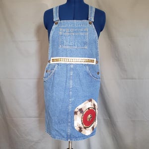 May include: Light blue denim overall dress with adjustable straps, a front pocket, and side pockets. The dress features a decorative white band with gold-colored accents and a circular patch with a red center and a cream-colored border.
