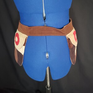 May include: A brown suede belt with two attached pouches. The pouches have a tan corduroy base with a red circle design and brown and tan leather accents. The belt is displayed on a blue dress form.