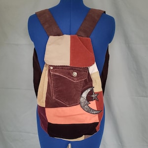 May include: A patchwork backpack with a variety of colors including brown, beige, red, and orange. The backpack has a front pocket with a button closure and a decorative crescent moon and star. The straps are brown.