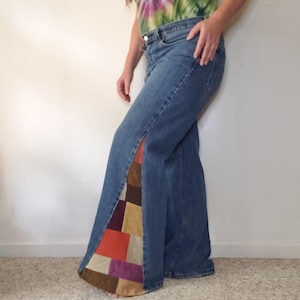 May include: Blue denim jeans featuring a side slit that reveals a multi-colored patchwork panel made of suede-like fabric squares in shades of brown, orange, purple, and tan. The jeans have a classic waistband with a button and zipper closure.
