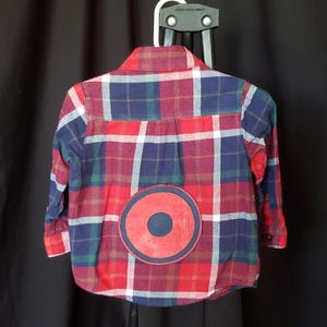 May include: A red, blue, and white plaid button-down shirt with a large red circle and a smaller blue circle applique on the back. The shirt is hanging on a white hanger against a black background.