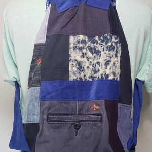 Może przedstawiać: Plecak ze sznurkiem typu patchwork wykonany z r&oacute;żnych niebieskich i szarych tekstur tkanin, w tym dżinsu, sztruksu i wzoru cętkowanego. Torba posiada wyraźną tylną kieszeń z zapięciem na guzik oraz wyhaftowane pomarańczowe logo kotwicy.