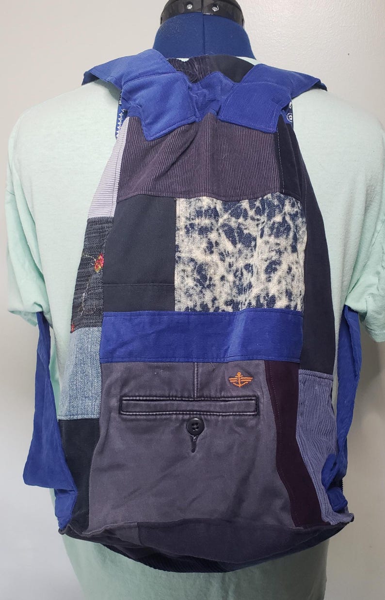 Może przedstawiać: Plecak ze sznurkiem typu patchwork wykonany z r&oacute;żnych niebieskich i szarych tekstur tkanin, w tym dżinsu, sztruksu i wzoru cętkowanego. Torba posiada wyraźną tylną kieszeń z zapięciem na guzik oraz wyhaftowane pomarańczowe logo kotwicy.