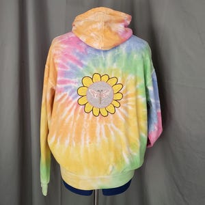 May include: A tie-dye hoodie in a rainbow of colors, including yellow, orange, pink, blue, and green. The back of the hoodie features a sunflower design with a butterfly in the center. The hoodie has a hood and long sleeves.