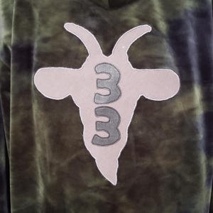 Pode incluir: Moletom tie-dye verde e preto com um logotipo de cabe&ccedil;a de cabra nas costas. O logotipo &eacute; de cor bege claro com o n&uacute;mero "33" em cinza. O moletom tem capuz e uma textura macia e aveludada.