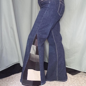 May include: Dark wash denim bell-bottom jeans with a patchwork design on the outer seams. The patchwork features squares of various neutral colors, including black, gray, and beige. The jeans have a classic five-pocket design.