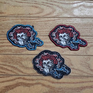 May include: Three embroidered patches featuring a skull with a floral crown. The patches have a gray background with red roses and are outlined in blue, red, and black thread. The patches are on a wooden surface.