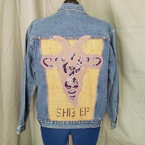 May include: Light blue denim jacket with a large goat head design on the back. The goat head is a light brown color with purple accents and a floral pattern. The text "SHIB EP" is printed below the design.