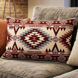 Pode incluir: Almofada decorativa retangular com fundo creme e um padrão geométrico em tons de bordô, azul claro e creme. A almofada está colocada em um sofá cinza com outras almofadas.