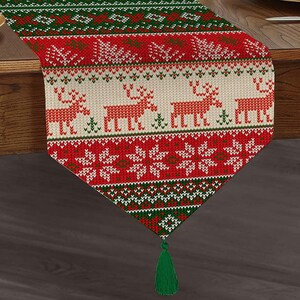 Puede incluir: Un camino de mesa de Navidad rojo, verde y blanco con un patrón tejido que presenta renos y copos de nieve. El camino de mesa tiene una borla verde en el extremo.